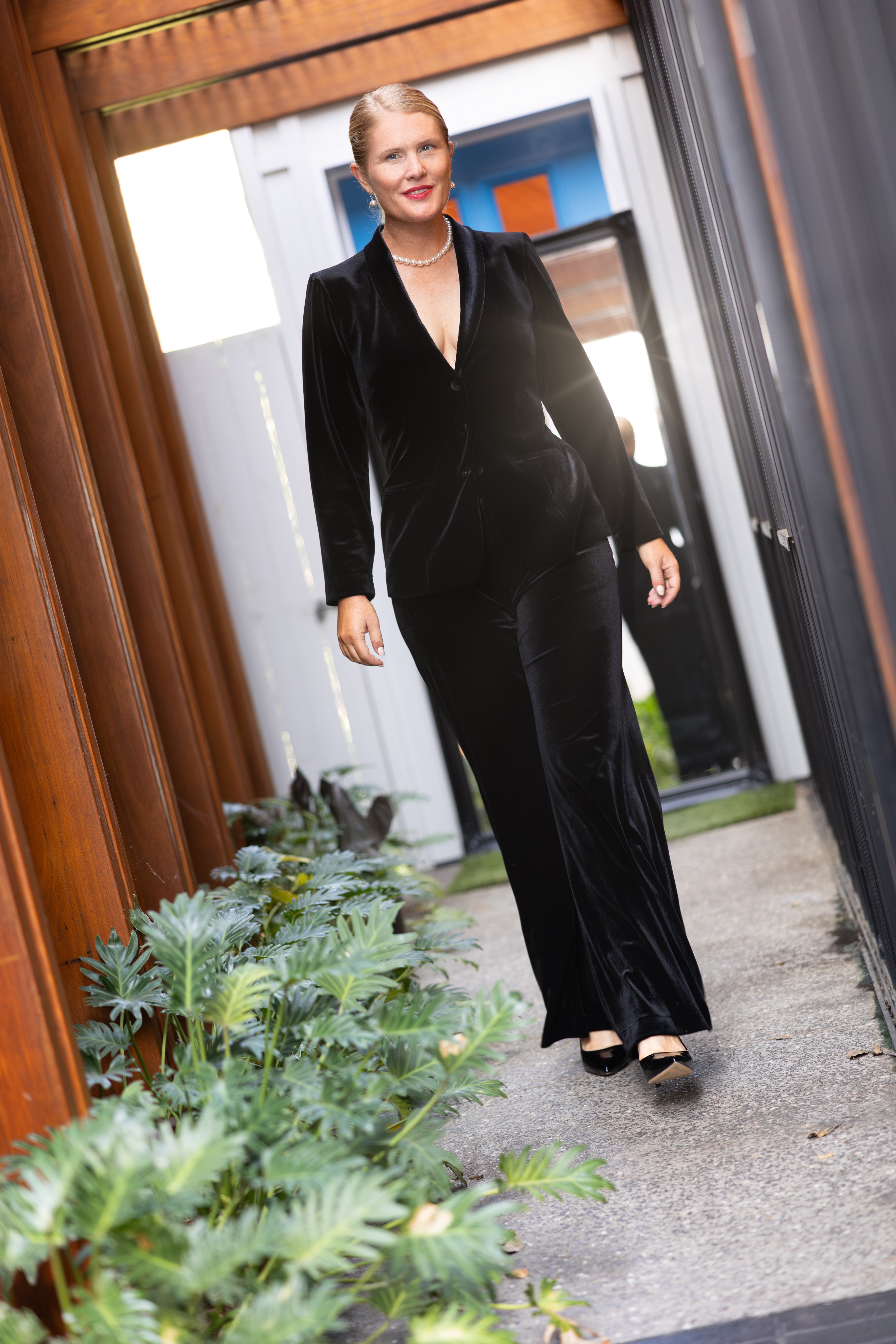 Woman in a black velvet coat and pants standing outdoors near a building entrance.