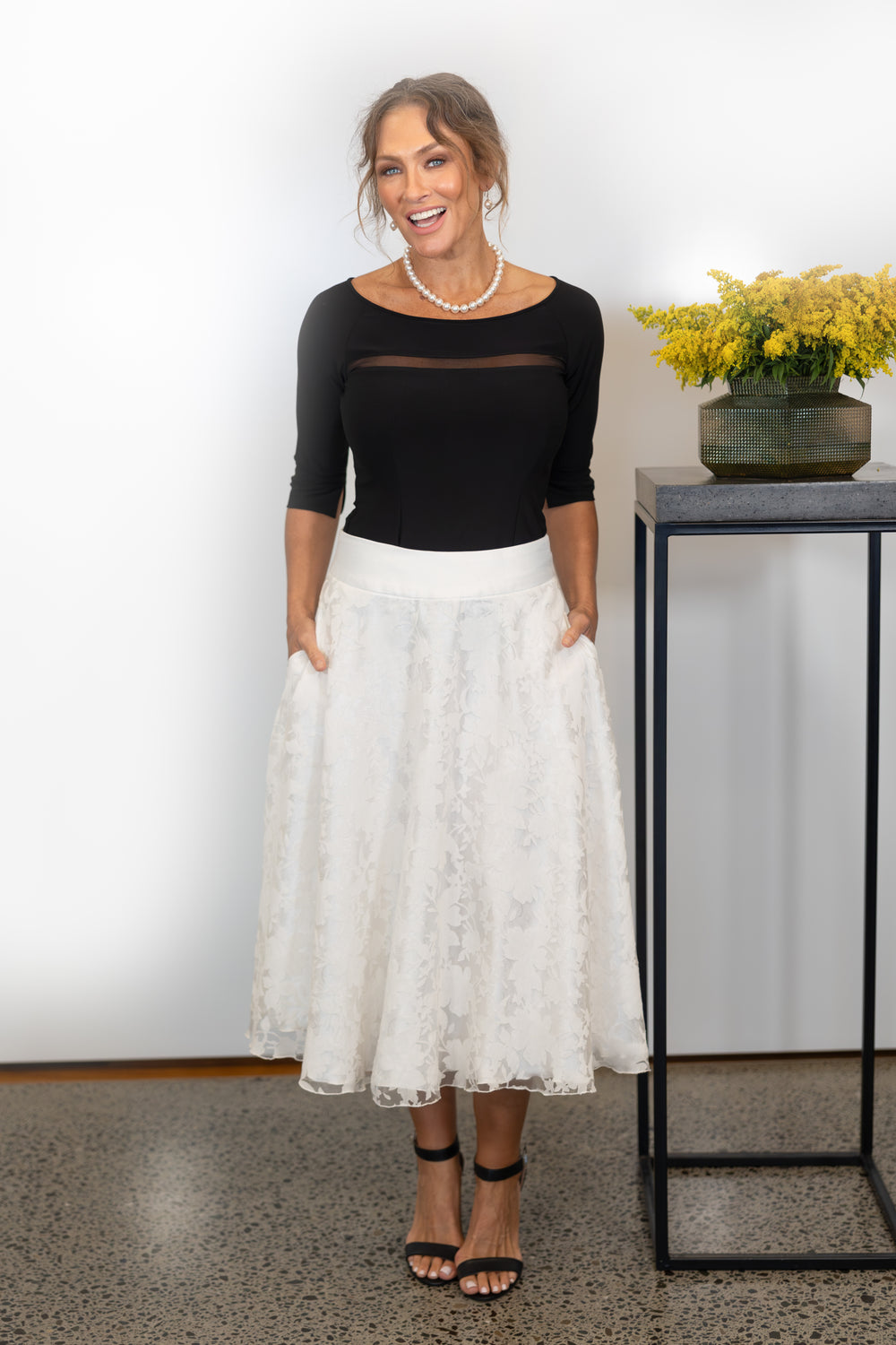 Woman wearing a black top and white lace skirt standing indoors with a decorative table and flowers in the background.